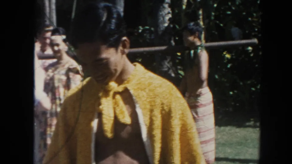 The performer in a yellow cape looks downward and smiles in 1961 Hawaii footage, with spectators, railing, and greenery behind him.