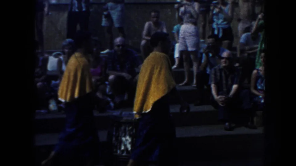 1961 Hawaii 8mm frame showing two performers in yellow capes and dark wraps before a seated crowd outdoors.
