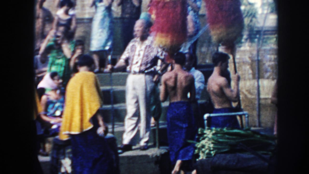 A man stands near performers and spectators during an outdoor Hawaiian gathering.