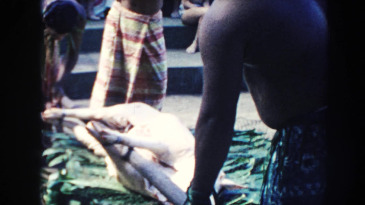 Pig laid on green leaves while people in patterned wrap skirts stand nearby.