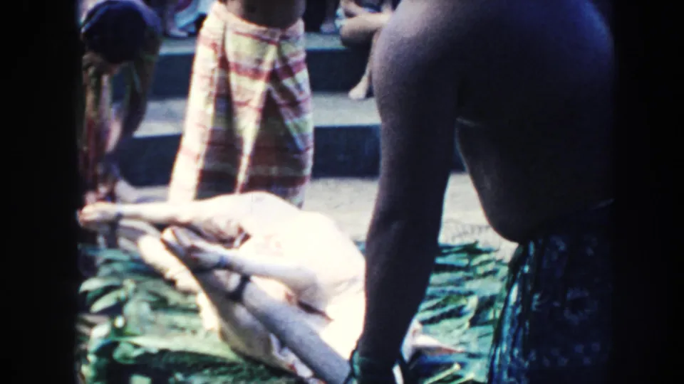 1961 Hawaii home-movie frame showing a pig on green leaves as people in patterned wrap skirts gather around it.