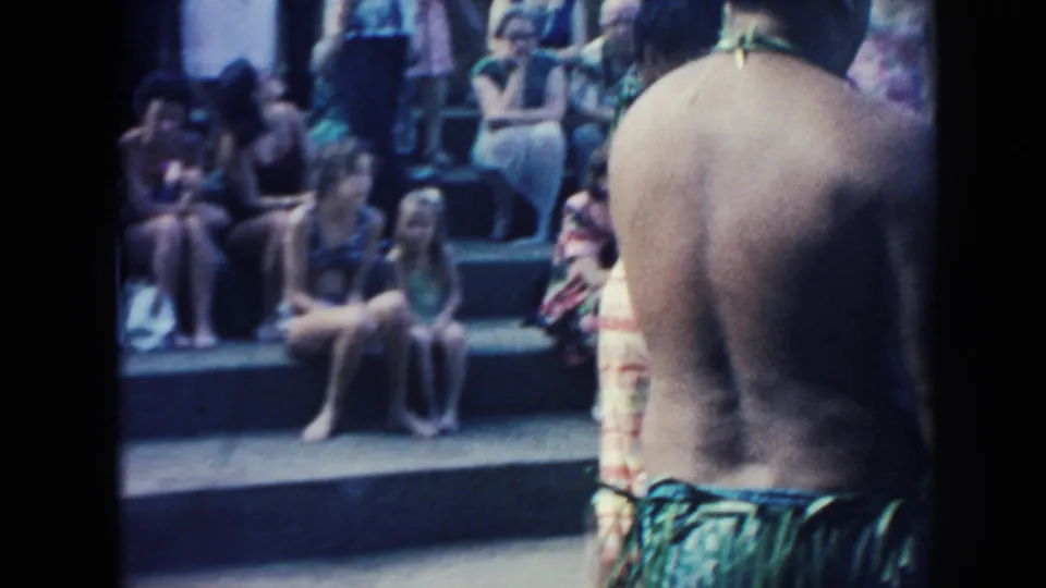 1961 Hawaii home-movie view of a shirtless man in a patterned wrap standing before seated spectators at a luau-style event.