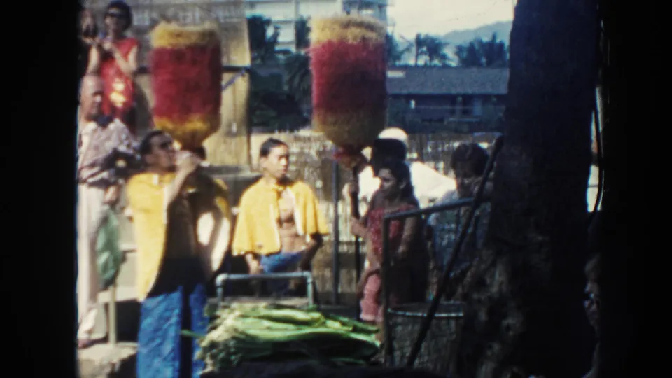 1961 Hawaii luau frame showing people near red and yellow feather-like standards with green leaves and railings in the foreground.