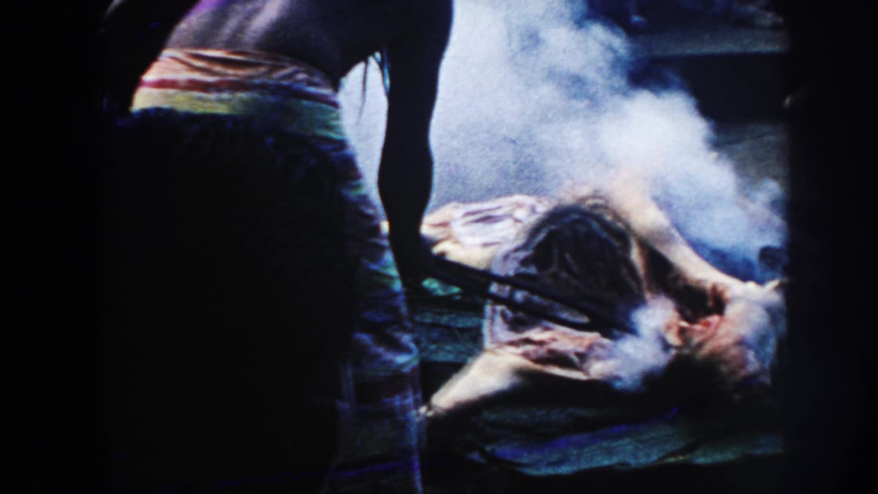 1961 Hawaii 8mm frame showing a shirtless person in a colorful wrap beside smoking meat on a bed of leaves.