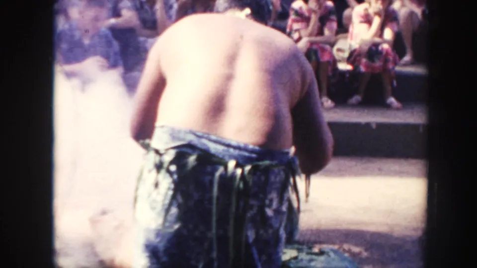 1961 Hawaii footage shows a shirtless man from behind near drifting smoke, with seated spectators in the background.
