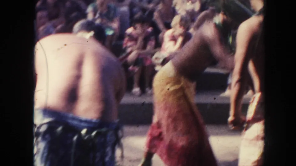 A 1961 Hawaii 8mm frame shows men in wrap garments near smoke as spectators sit on stepped seating behind them.