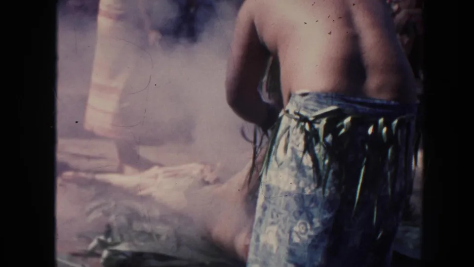 In 1961 Hawaii footage, a barefoot performer in a patterned wrap stands close to smoke and dark coals during an outdoor act.