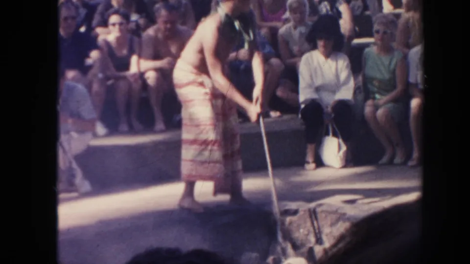 The performer bends forward with long tongs near the coal area while seated spectators watch in the 1961 Hawaii home movie.