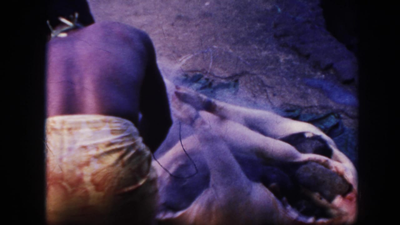 1961 Hawaii 8mm frame of a shirtless man in a patterned wrap beside a large animal carcass on bare ground.
