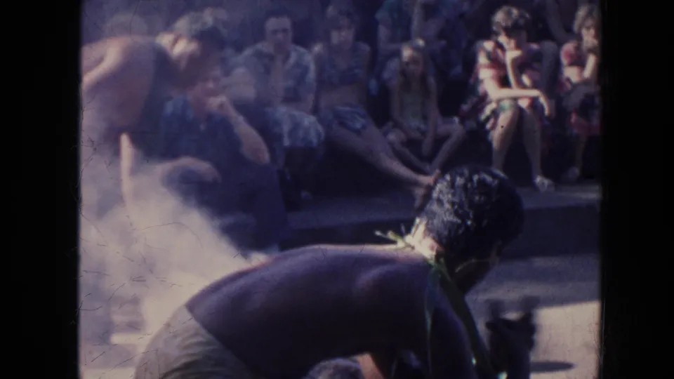 1961 Hawaii 8mm frame showing adults around a smoky outdoor scene with seated onlookers in the background.