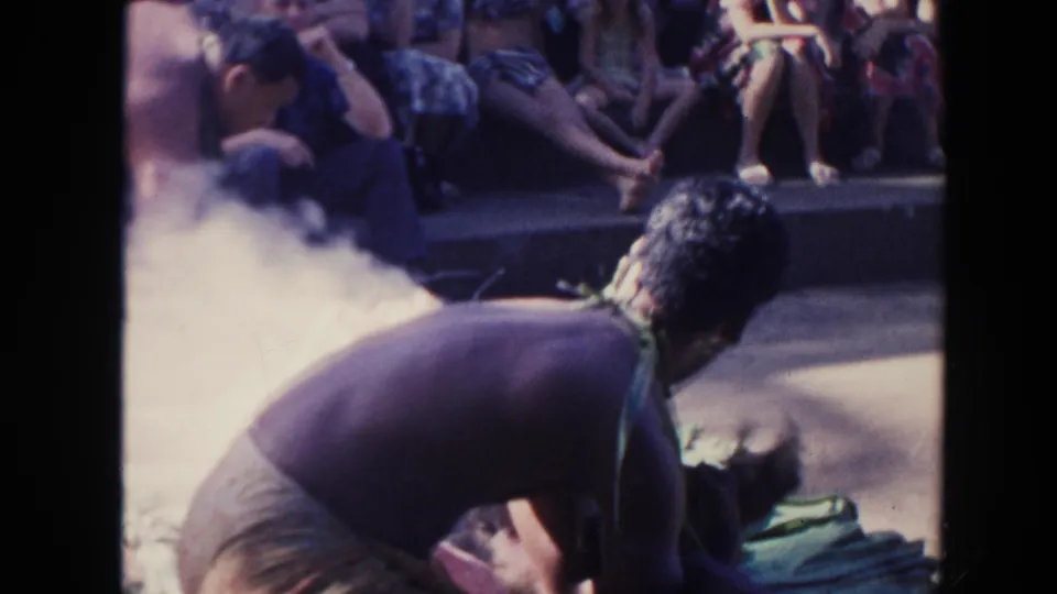 1961 Hawaii home-movie frame of a shirtless adult leaning near smoke with spectators seated behind.