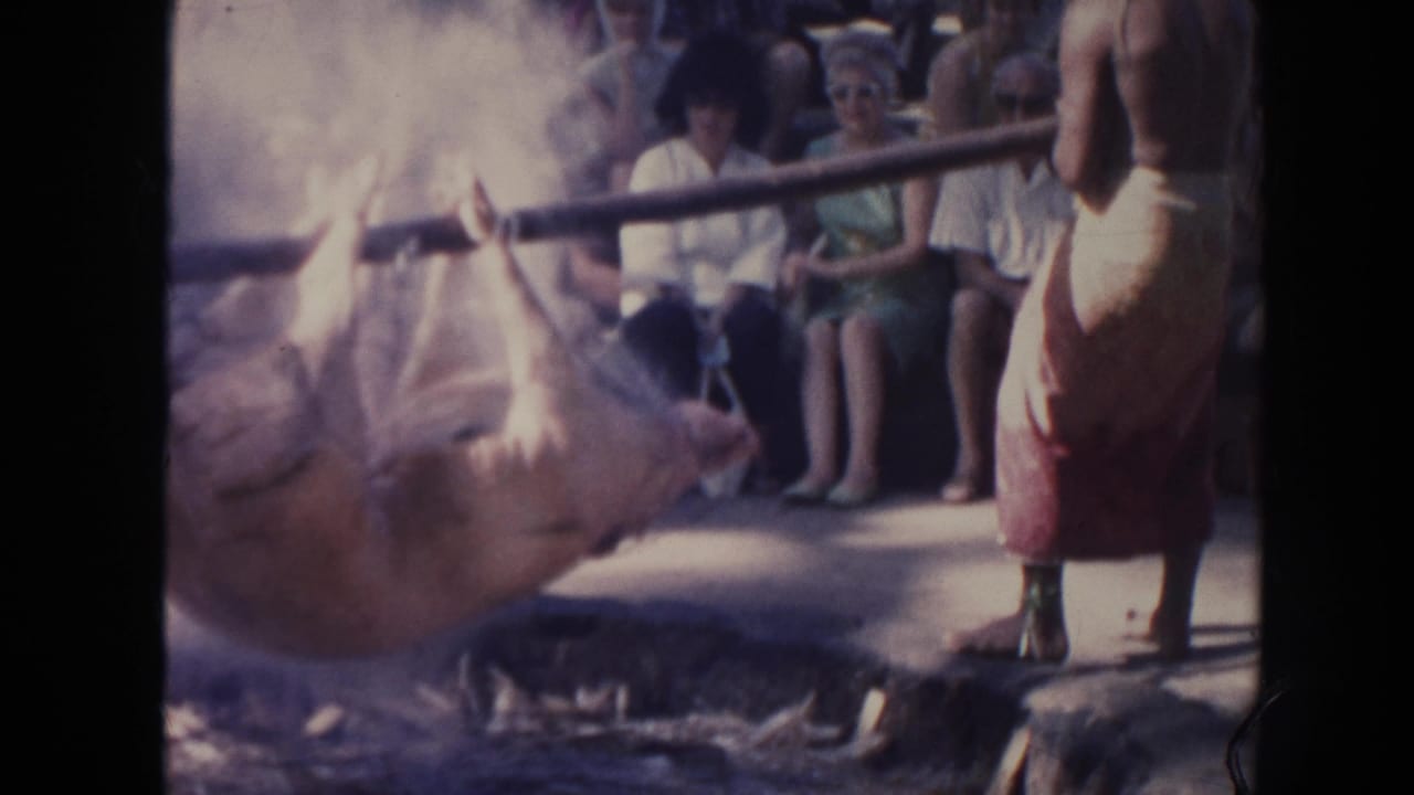 1961 Hawaii 8mm frame showing a pig roasting over smoke while seated and standing spectators watch nearby.