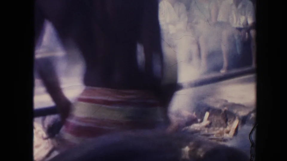 A blurred figure passes close to the camera during a smoky 1961 Hawaii pig roast scene with spectators beyond.