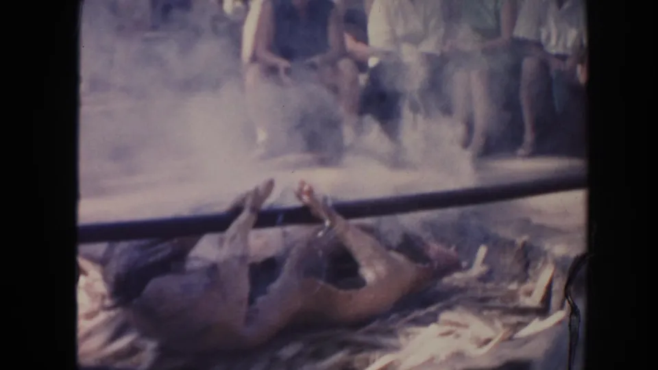 Late frame of the Hawaii pig roast, with the animal near the smoky pit and adults gathered behind a low railing.