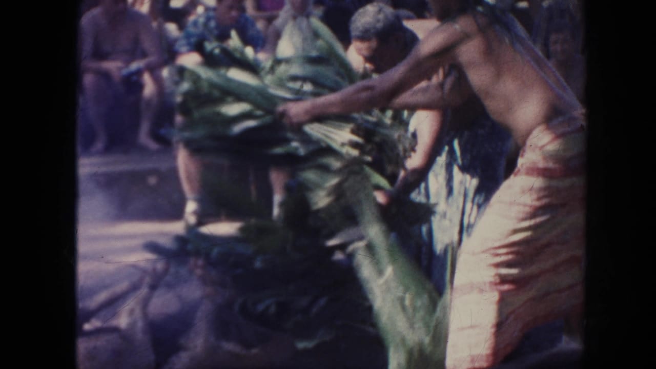 1961 Hawaii luau scene with a man in a patterned wrap moving large green leaves as seated spectators watch.