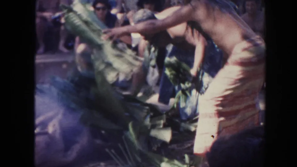 Handheld 8mm view of a man in a patterned wrap shifting broad leaves in a smoky 1961 Hawaii luau setting.