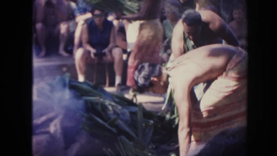 1961 Hawaii luau frame with men bending near the smoky pit and broad leaves while seated onlookers watch.