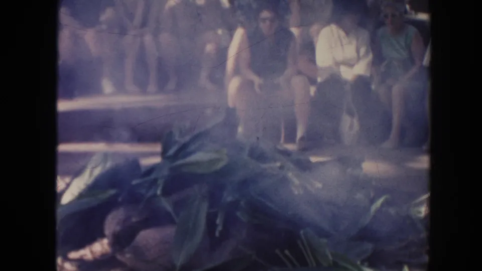 A smoky 1961 Hawaii 8mm frame shows large leaves in the foreground with spectators partly obscured behind them.