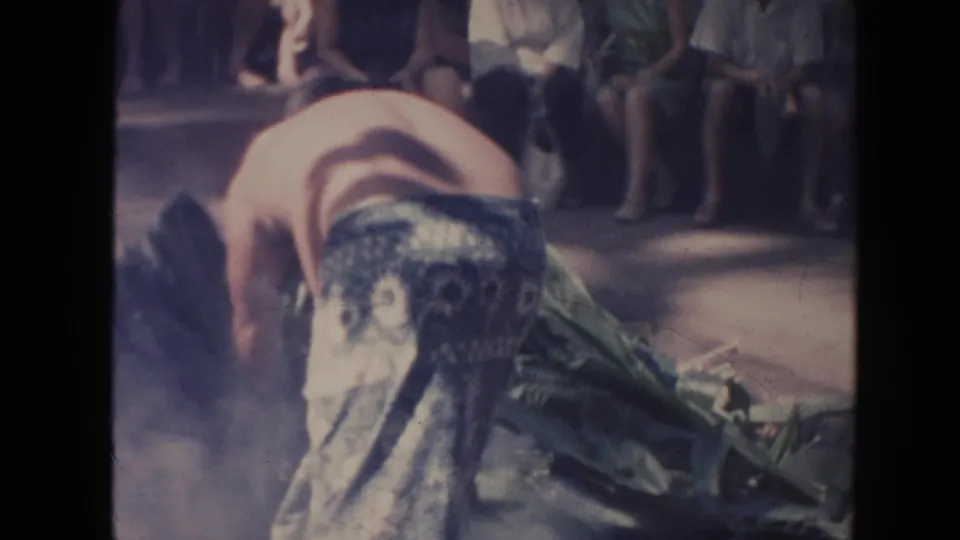 1961 Hawaii home-movie frame of a shirtless man bending over green leaves near smoke with seated spectators behind him.