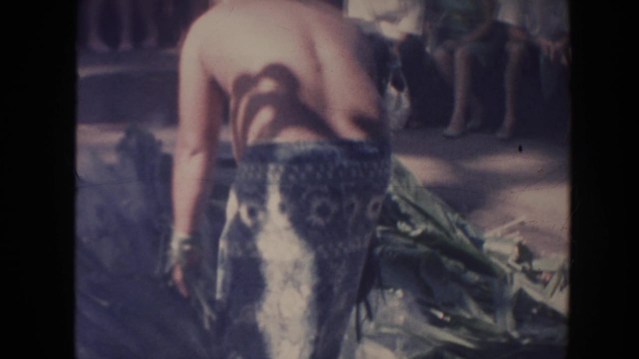 In 1961 Hawaii home-movie footage, a shirtless man in patterned shorts bends near green leaves while spectators stand or sit in the background.