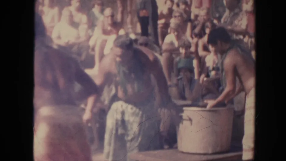 Two shirtless men in patterned clothing tend a smoky leaf fire or container area in Hawaii, while a seated crowd watches in the background.