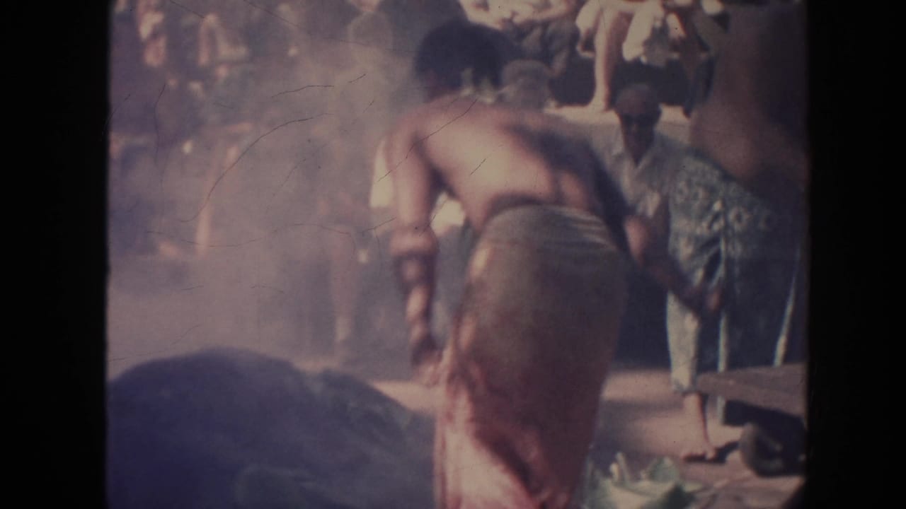 1961 Hawaii 8mm frame showing a performer in a wraparound skirt bending near a mat with spectators in the background.