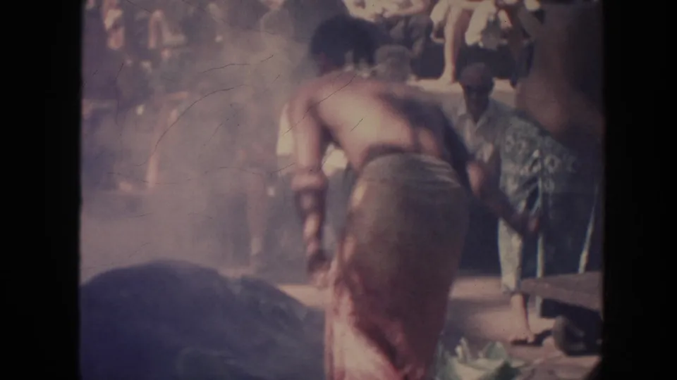 1961 Hawaii 8mm frame showing a performer in a wraparound skirt bending near a mat with spectators in the background.