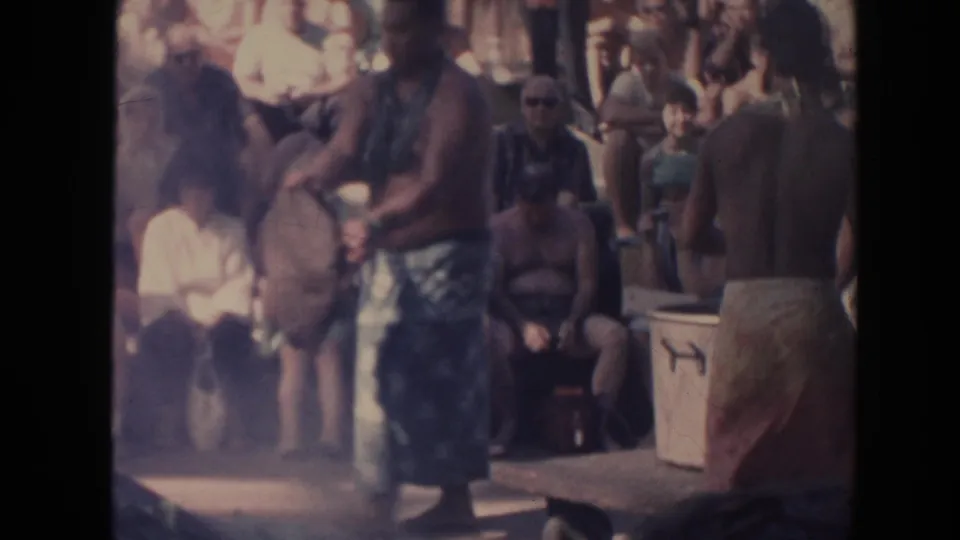 1961 Hawaii 8mm frame of performers gathered near a mat or rug with a seated audience close behind them.