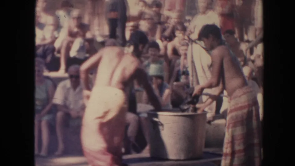 1961 Hawaii 8mm frame showing two performers near a large container and mat with spectators watching behind them.