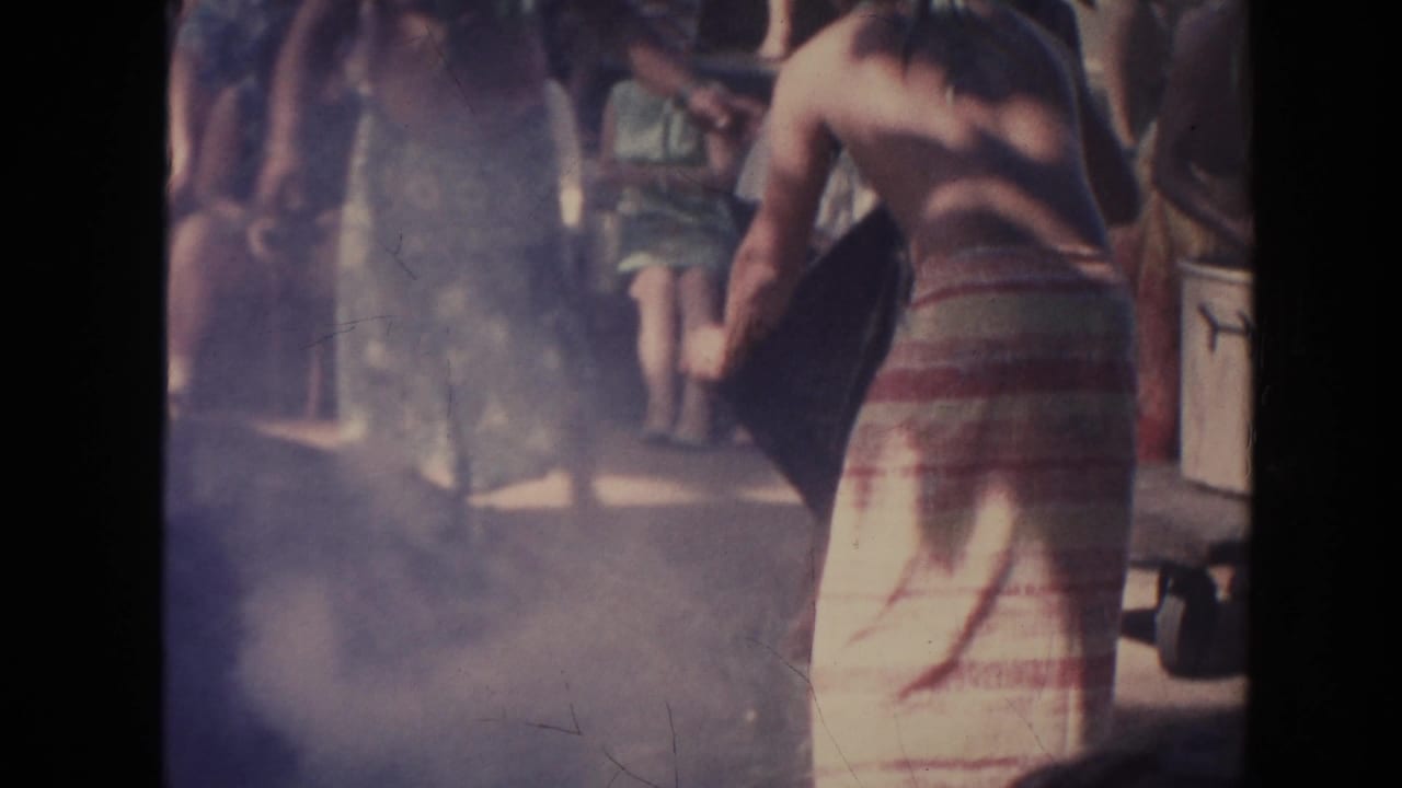 Shirtless man in a striped wrap tends a smoky area while spectators stand nearby.