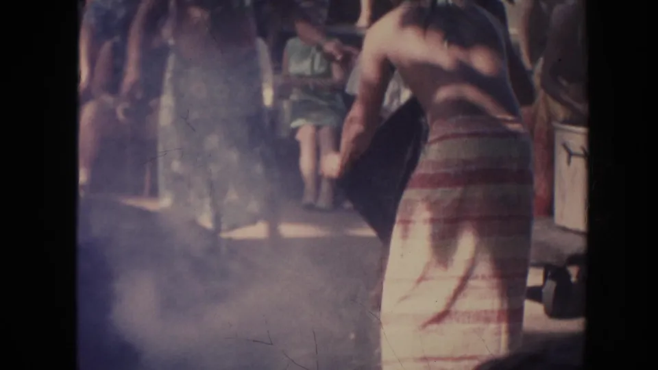 1961 Hawaii 8mm frame showing a shirtless man in a striped wrap near smoke or haze with onlookers gathered behind him.
