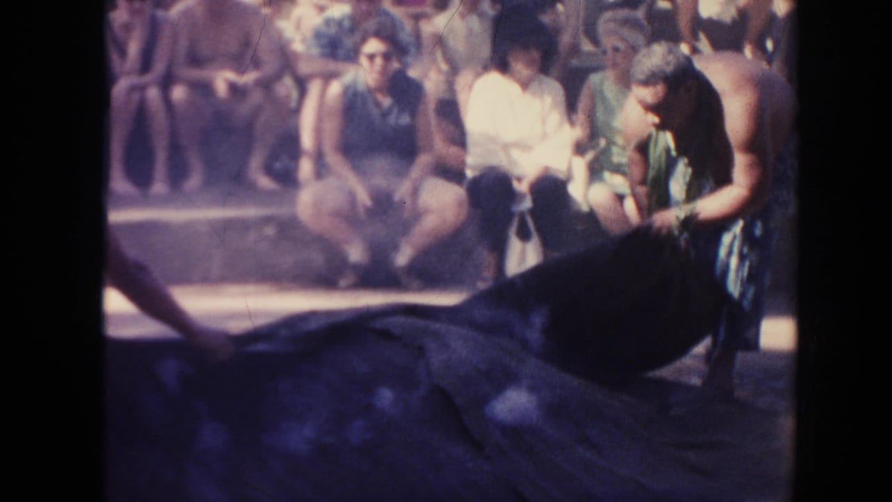 Man bends near a large dark covering while seated spectators watch from steps.