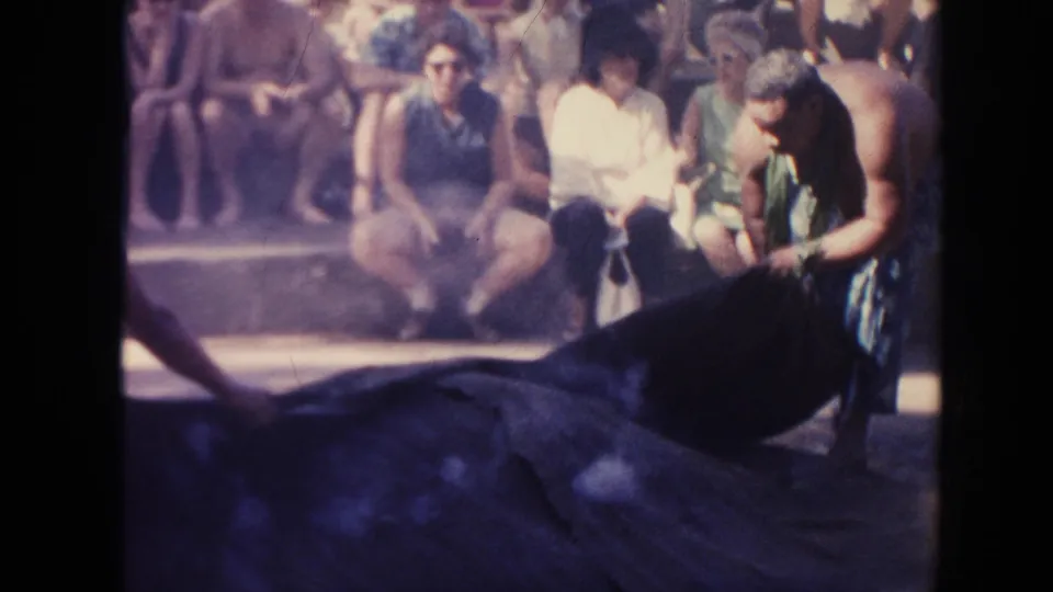 1961 Hawaii home-movie frame with a man leaning over a large dark ground covering as spectators sit on steps behind him.