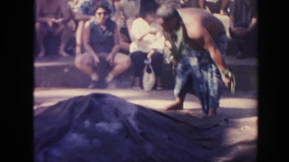 1961 Hawaii 8mm frame showing a man in a wrap skirt reaching toward a dark tarp or blanket with spectators in the background.