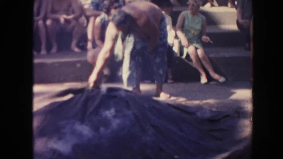 1961 Hawaii frame of a shirtless man adjusting a broad dark covering on the ground while visitors or spectators watch from steps.