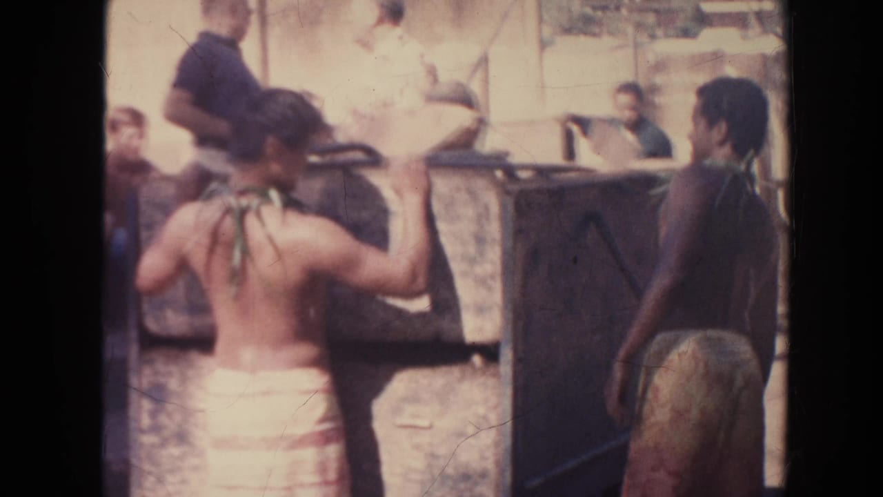 Shirtless men in wraparound skirts stand beside a large wooden wagon outdoors.