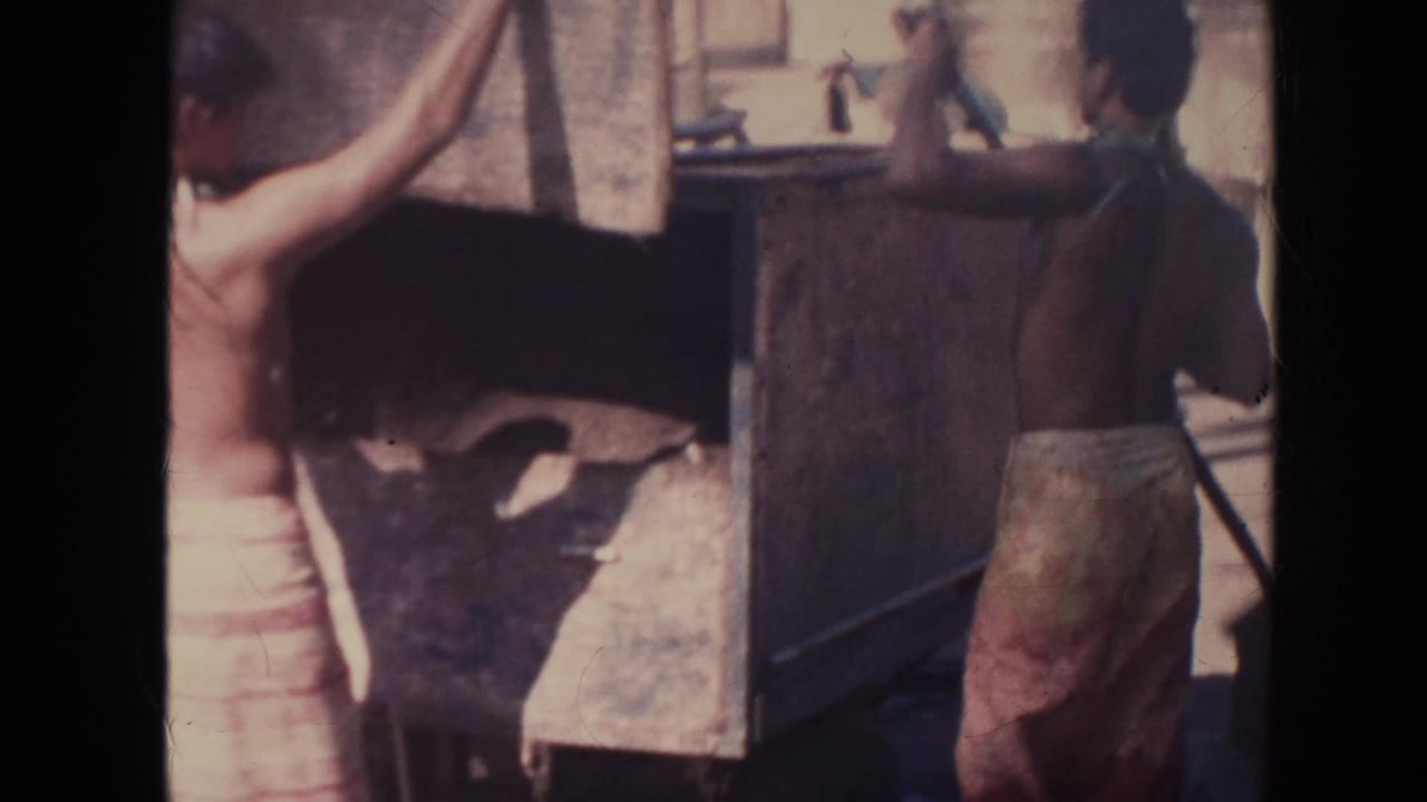 Two shirtless men in wraparound skirts appear to handle or steady a large wooden wagon in 1961 Hawaii footage.