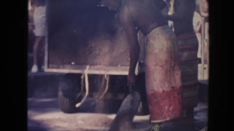 A shirtless man in a wraparound skirt stands next to the wooden wagon, with a sack or tool visible near the lower frame.