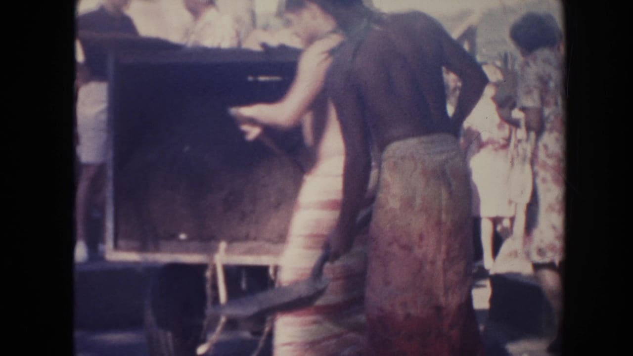 Men in wraparound skirts shovel dirt near a large container outdoors.