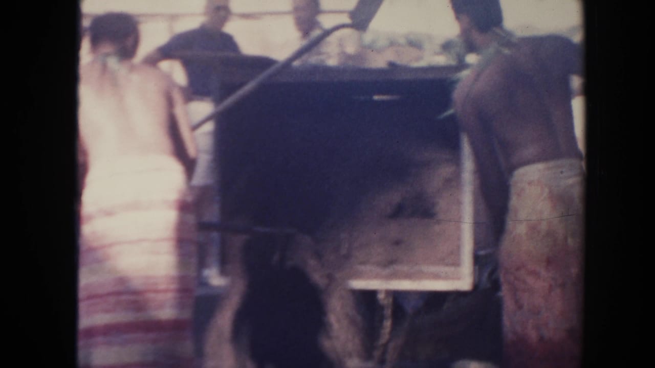 Workers stand beside a large dark container with a long shovel or pole visible.