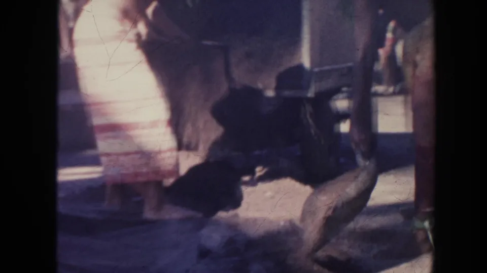 1961 Hawaii home-movie frame with a bending worker, a large container, and a woman in a light wraparound skirt nearby.