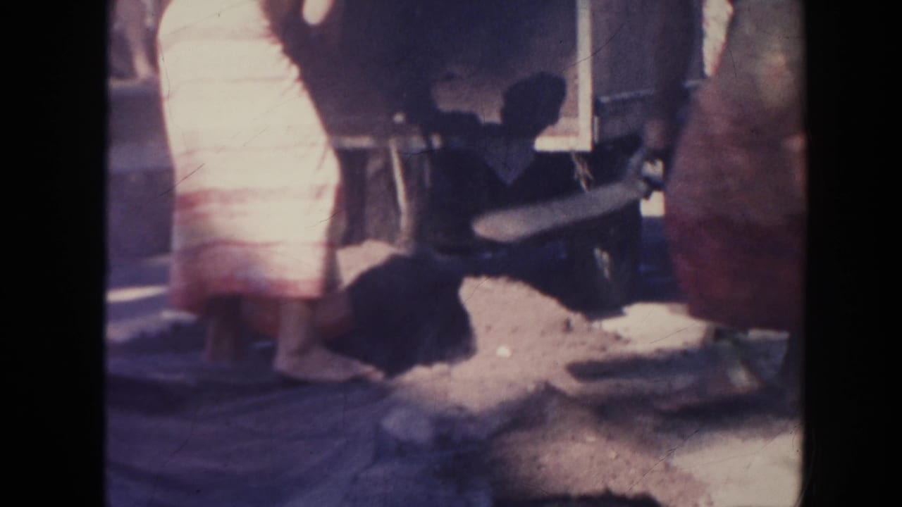 People stand near a pit as a man bends down with a shovel in bright outdoor light.