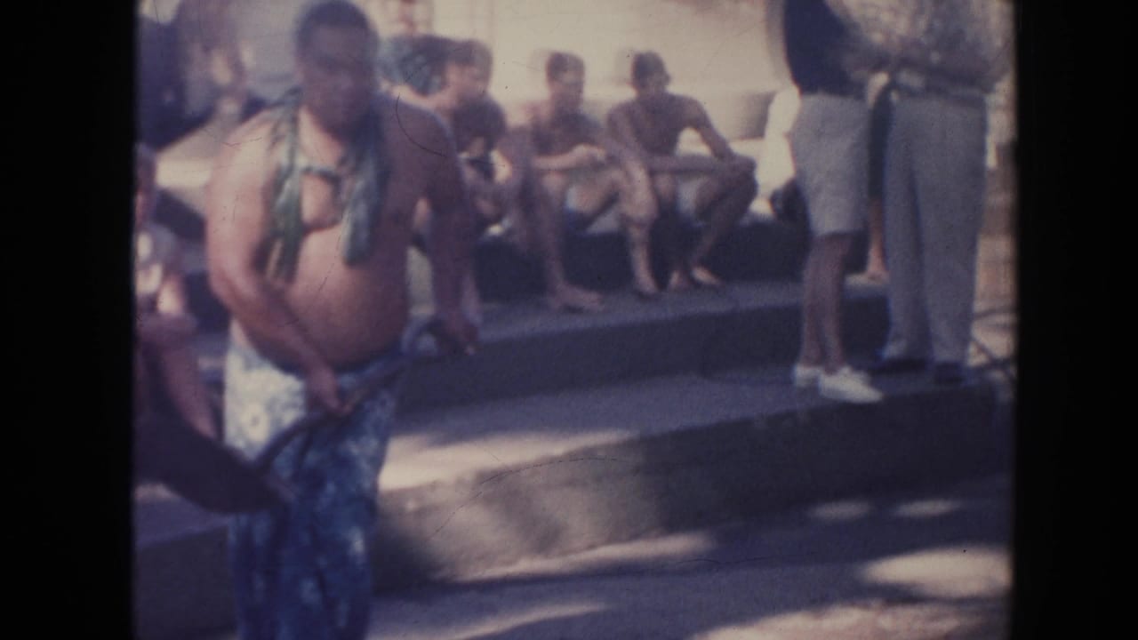 1961 Hawaii frame with a man in patterned wrap clothing and leafy adornment standing near steps where several people sit watching.