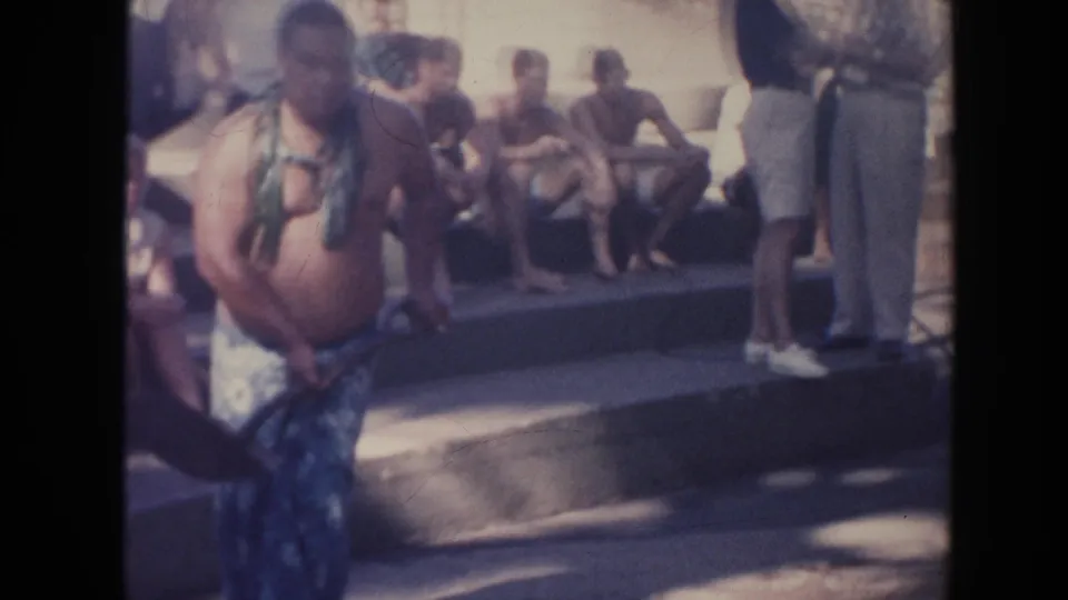 1961 Hawaii frame with a man in patterned wrap clothing and leafy adornment standing near steps where several people sit watching.