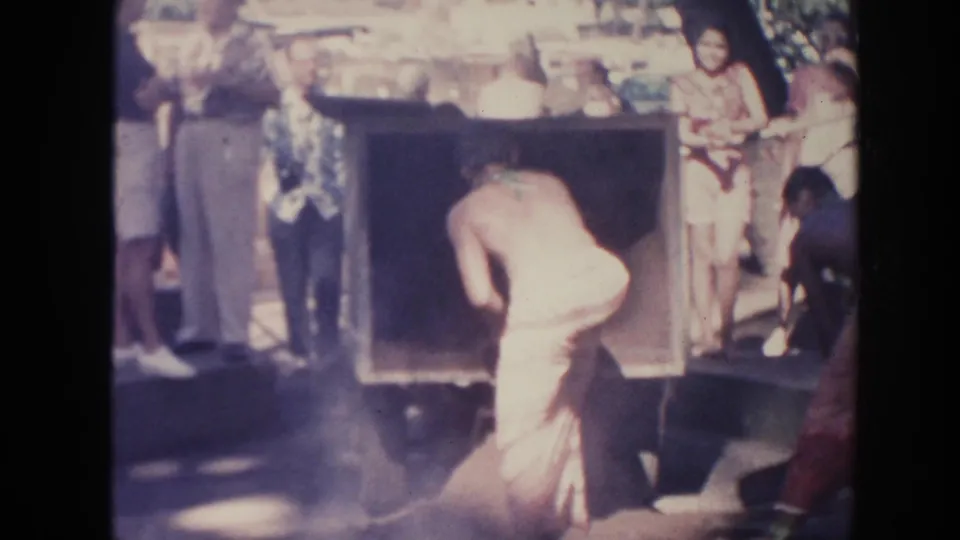 1961 Hawaii home-movie frame showing a man leaning into a rectangular pit or oven opening as nearby adults observe.