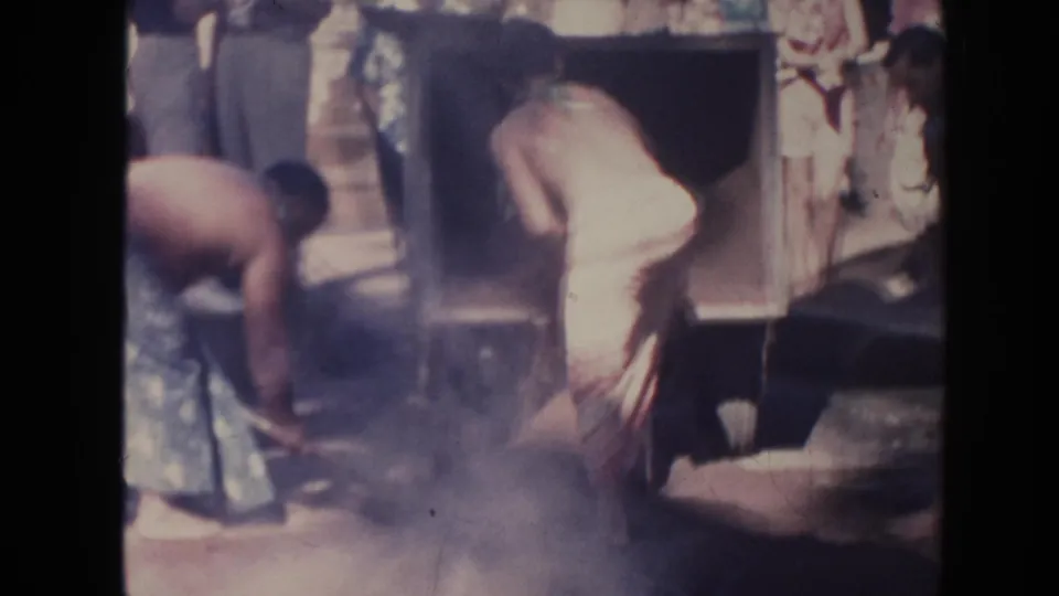 1961 Hawaii frame showing men in wrap garments working beside a pit as haze or dust rises around seated and standing onlookers.