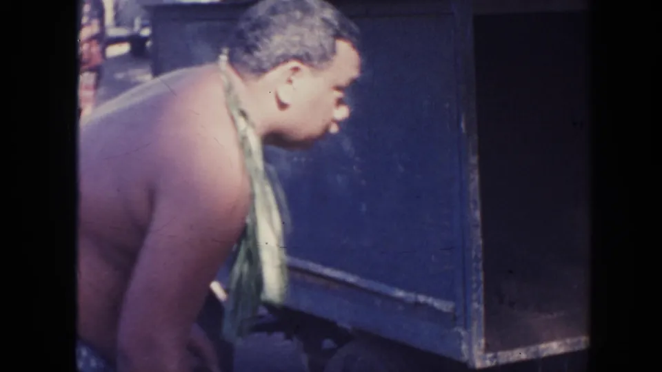 1961 Hawaii 8mm frame showing a shirtless man leaning near a blue metal container, with other figures in the background.