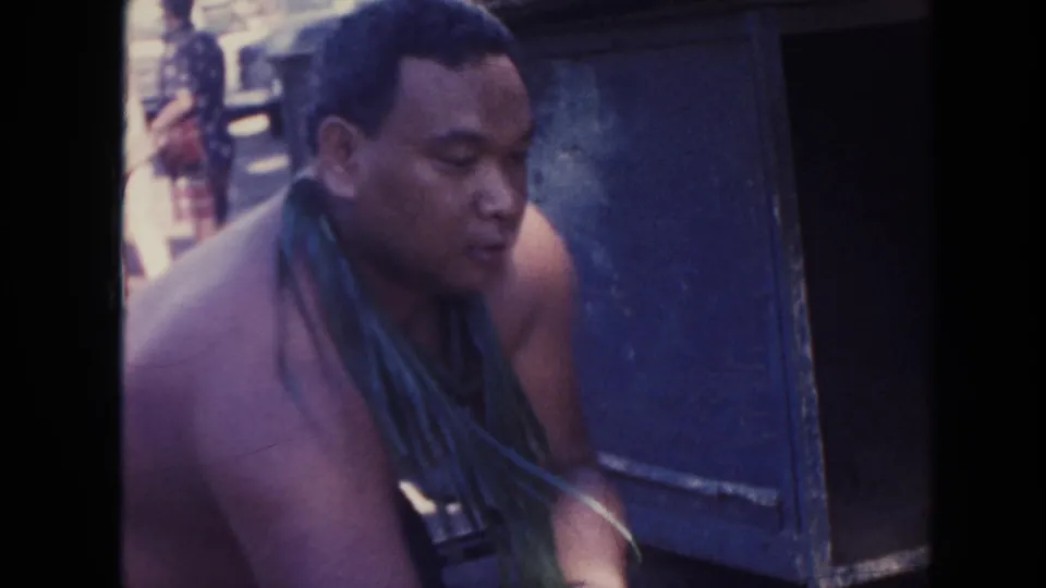 1961 Hawaii 8mm close frame of a shirtless man wearing a green cloth or wrap beside a blue metal container.