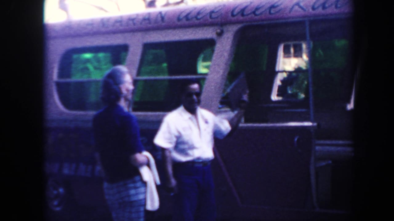 1961 Hawaii frame with a man holding a clipboard and gesturing near a vintage bus as a woman watches.