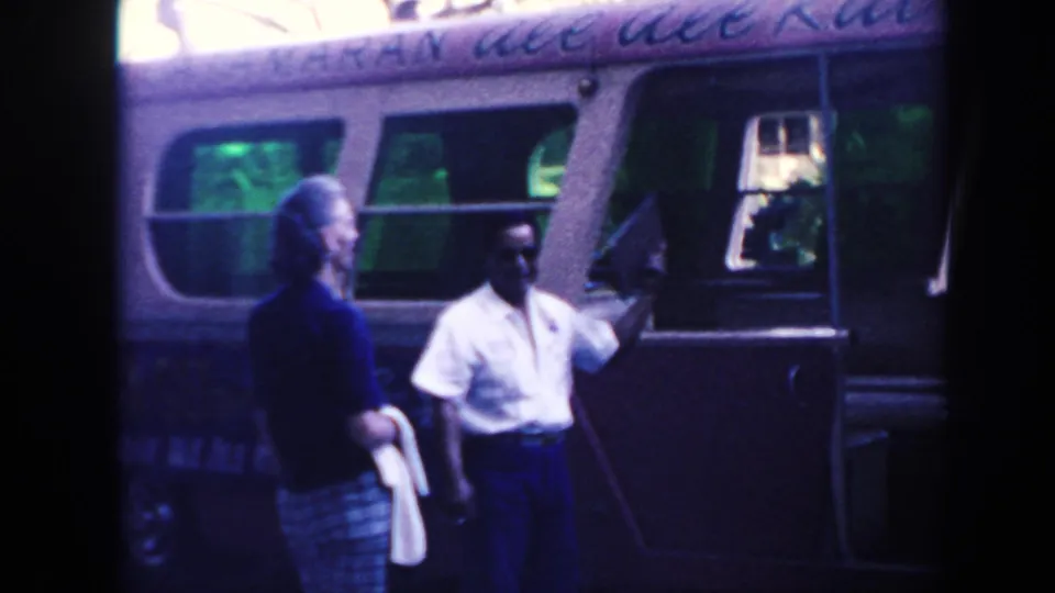 1961 Hawaii frame with a man holding a clipboard and gesturing near a vintage bus as a woman watches.